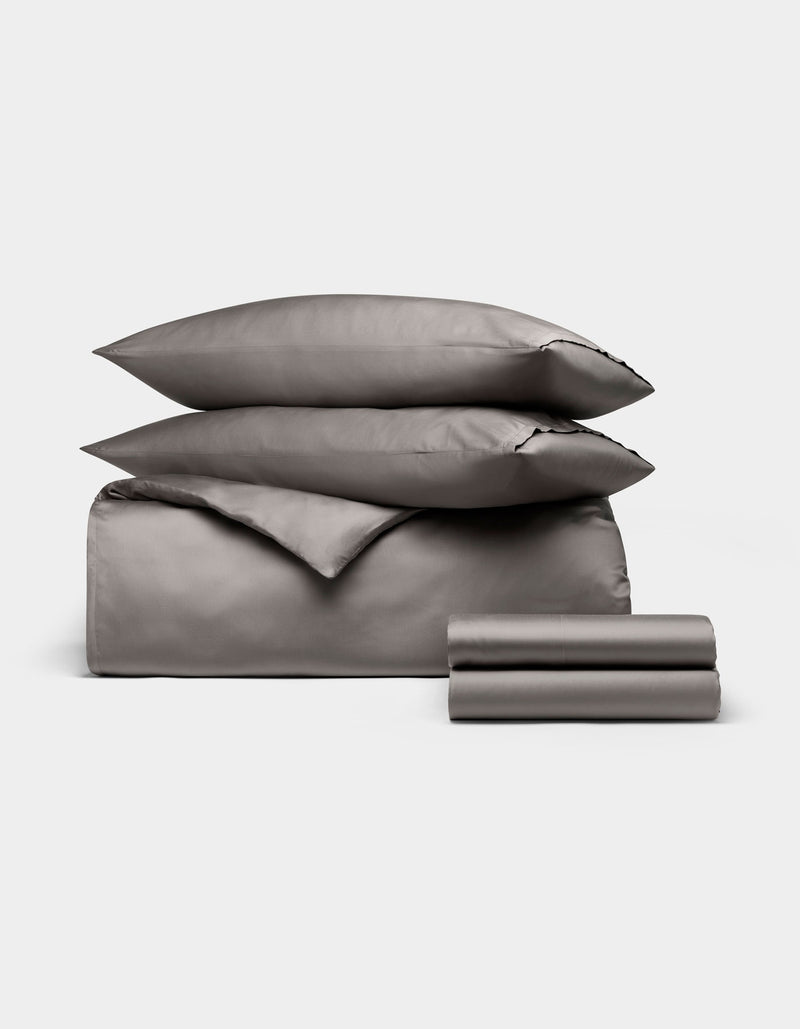 The Cozy Earth Bamboo Bedding Deluxe Bundle features neatly folded gray sheets, pillowcases, a duvet cover, flat and fitted sheets, and two stacked pillows displayed on a plain white background.