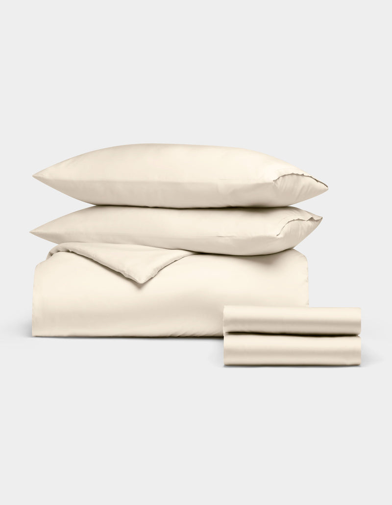 The Cozy Earth Bamboo Bedding Deluxe Bundle, featuring a cream comforter, two pillows, and two stacked sheets, is neatly folded and displayed against a plain light gray background.