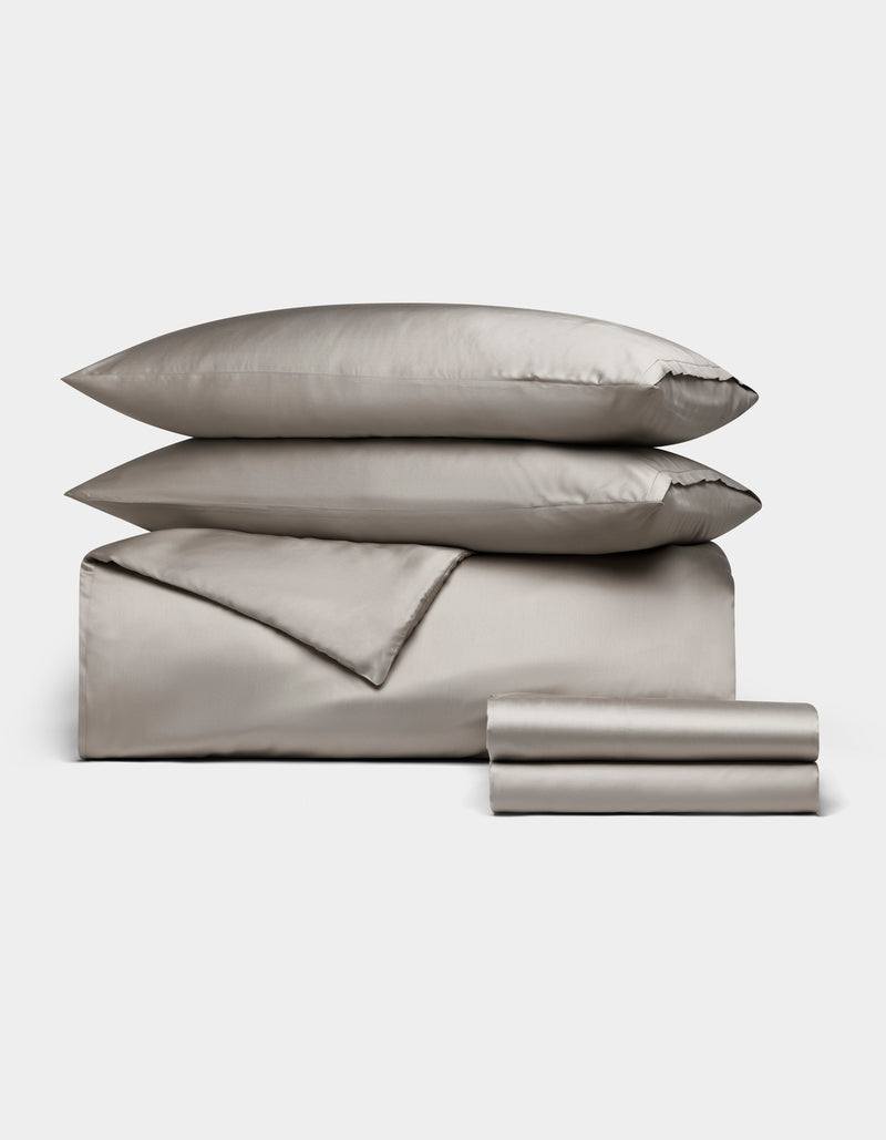 The Bamboo Bedding Deluxe Bundle by Cozy Earth features a neatly arranged silver-gray set with two pillows, a folded duvet, and stacked sheets, displayed on a plain white background.