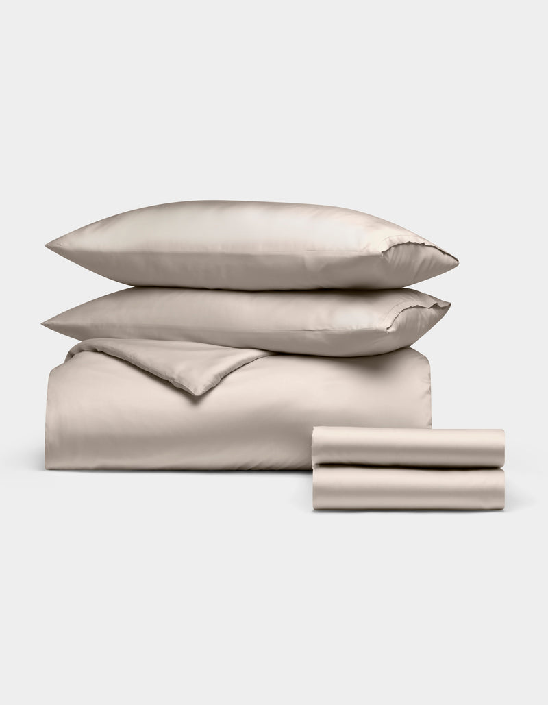 The Cozy Earth Bamboo Bedding Deluxe Bundle features two stacked pillows, a comforter, and folded sheets in cream, all neatly arranged against a plain white background.