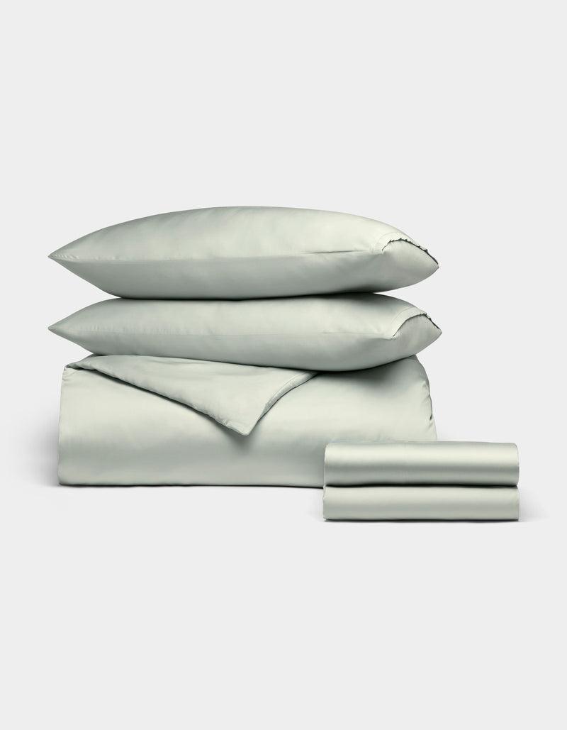 The Cozy Earth Bamboo Bedding Deluxe Bundle features a pale green set with two stacked pillows, a comforter, and folded sheets, elegantly displayed against a plain light background.