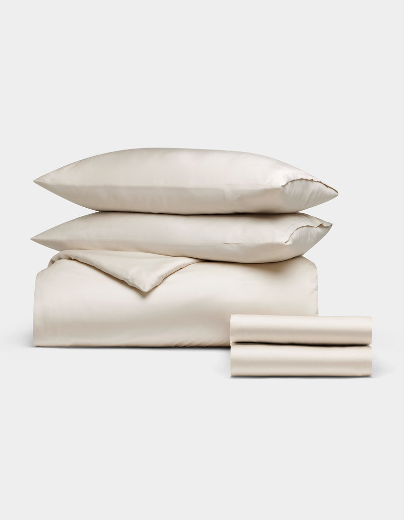Cozy Earth's Bamboo Bedding Deluxe Bundle, featuring two pillows, a folded comforter, and two folded sheets in cream, is neatly stacked and displayed on a plain white background.