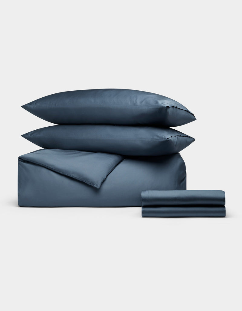 The Cozy Earth Bamboo Bedding Deluxe Bundle, featuring two pillows, a comforter, and folded sheets in blue, is neatly stacked against a plain light gray background.