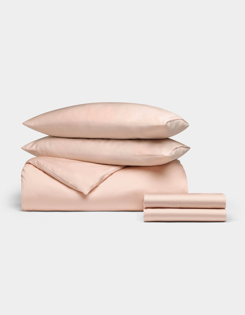 The Cozy Earth Bamboo Bedding Deluxe Bundle, featuring two pillows, a comforter, and two folded sheets in pale pink, is neatly stacked and displayed against a plain white background.