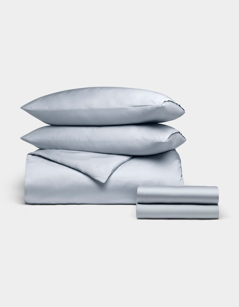 The Cozy Earth Bamboo Bedding Deluxe Bundle features a neatly folded light blue set with two pillowcases, a flat sheet, fitted sheet, and comforter, all arranged on a plain light background.