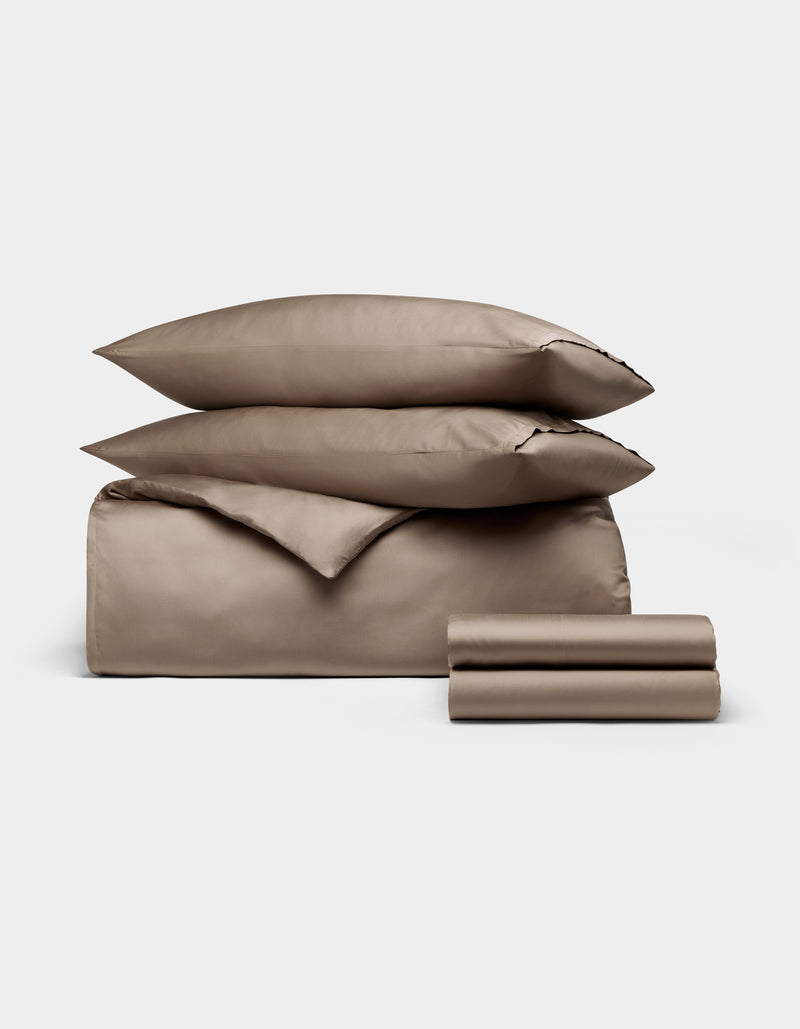 The Cozy Earth Bamboo Bedding Deluxe Bundle features taupe-colored bed linens with two stacked pillows, a folded duvet, and sheets, all neatly arranged against a plain white background.