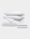 The Bamboo Bedding Deluxe Bundle by Cozy Earth features a neatly stacked set of white bedding—two pillows, a comforter, and a folded sheet—displayed against a plain light gray background.|Color:White