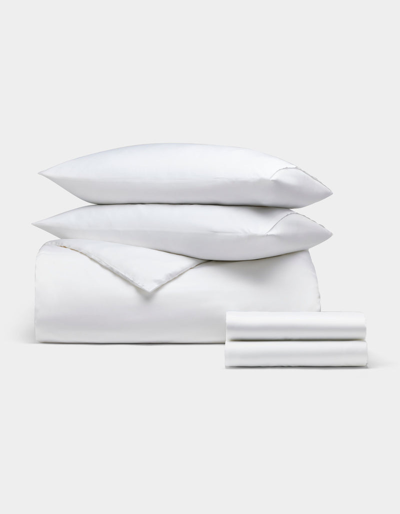 The Bamboo Bedding Deluxe Bundle by Cozy Earth features a neatly stacked set of white bedding—two pillows, a comforter, and a folded sheet—displayed against a plain light gray background.