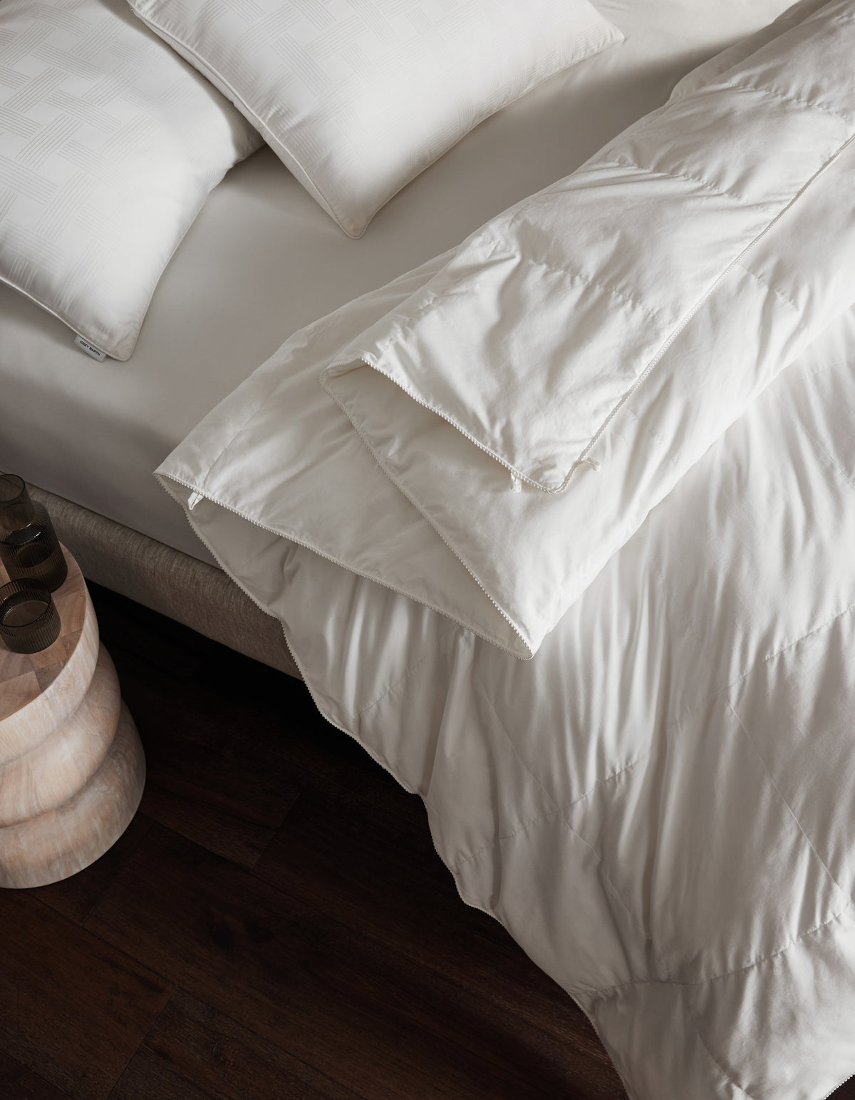 A neatly made bed features Cozy Earth's Down Alternative Comforter and white pillows beside a small wooden nightstand on a dark wood floor.|Filling:Lightweight