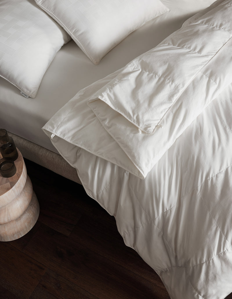 A neatly made bed features Cozy Earth's Down Alternative Comforter and white pillows beside a small wooden nightstand on a dark wood floor.