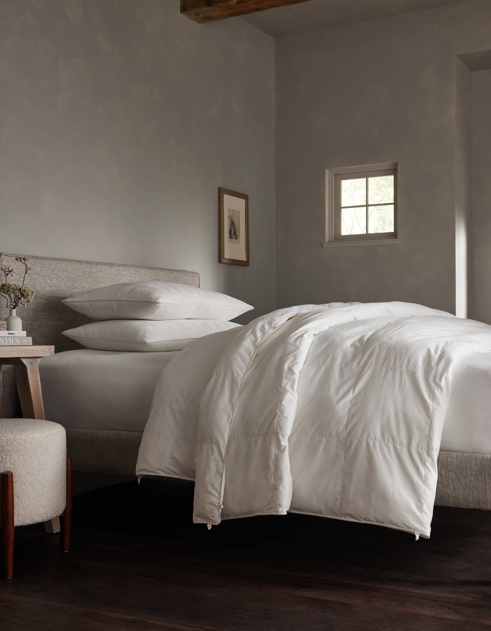 A modern bedroom with a neatly made bed featuring Cozy Earth’s Down Alternative Comforter, white bedding, a gray upholstered headboard, stacked pillows, framed wall art, a small window, and a round ottoman next to the wooden nightstand.