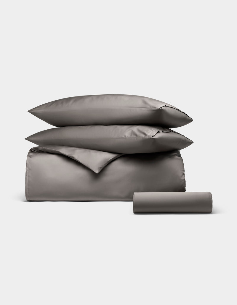 The Cozy Earth Bamboo Bedding Essential Bundle features a neatly stacked set of gray bedding—two pillows, a folded duvet, and a bolster pillow—displayed on a plain light background.