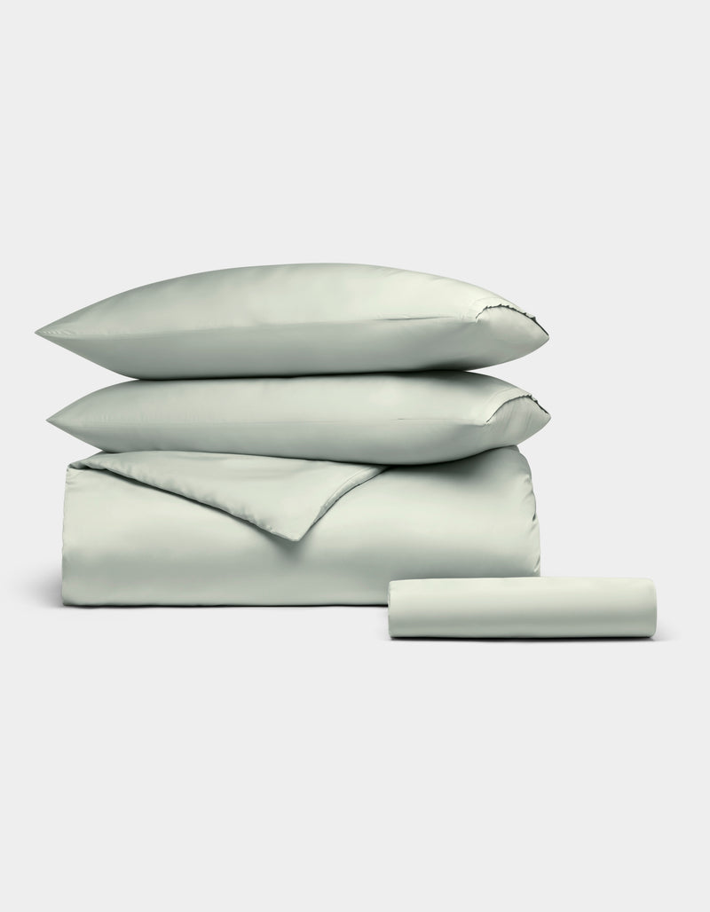 The Cozy Earth Bamboo Bedding Essential Bundle, featuring two pillows, a folded duvet, and a sheet in pale gray, is neatly stacked against a plain white background.