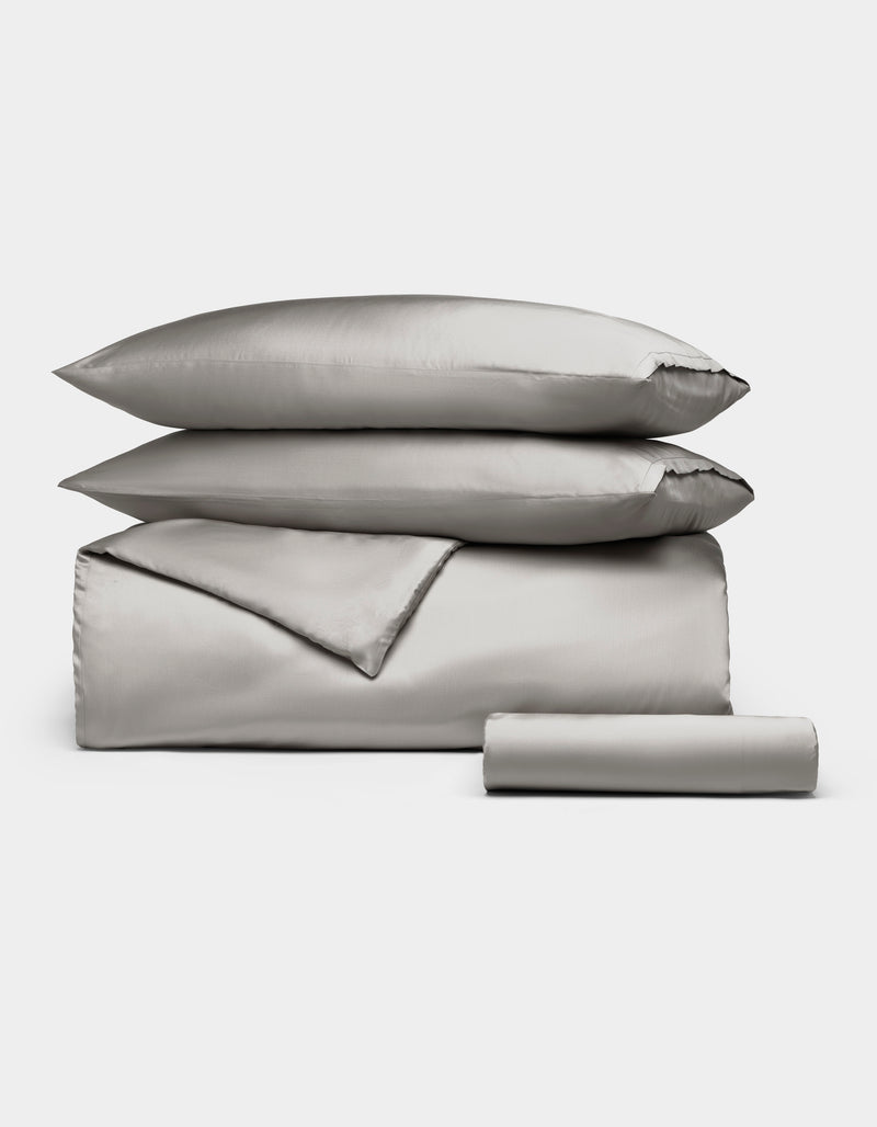 The Bamboo Bedding Essential Bundle by Cozy Earth in Light Grey is neatly arranged, featuring a pillowcase, duvet cover, flat sheet, and a rolled fitted sheet against a white background.