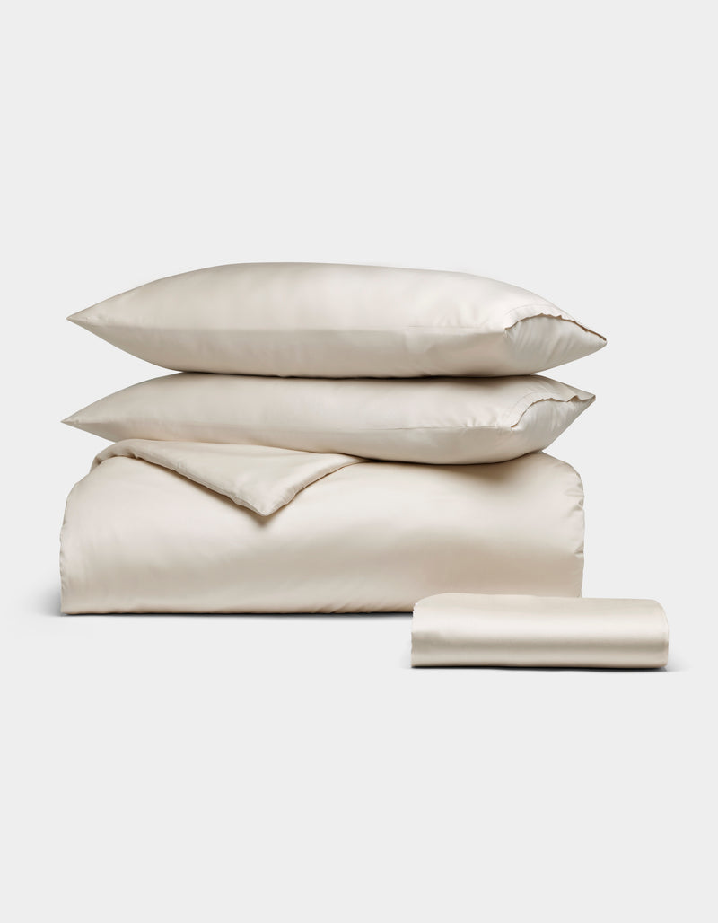 The Cozy Earth Bamboo Bedding Essential Bundle in oat, featuring two pillows, a folded duvet, and a folded sheet, is neatly stacked and displayed against a plain white background.