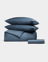 The Bamboo Bedding Essential Bundle by Cozy Earth features two pillows, a folded comforter, and a rolled bedsheet in blue, all neatly arranged against a plain white background.|Color:Pacific Blue