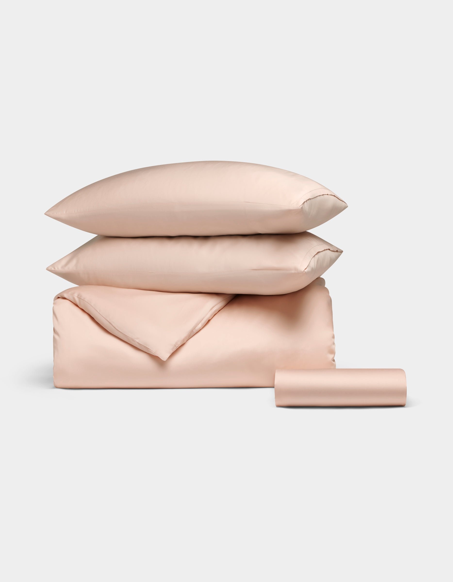The Bamboo Bedding Essential Bundle by Cozy Earth, featuring light pink satin pillows, a folded duvet, and a rolled sheet, is neatly arranged against a light grey background.