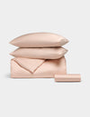 The Bamboo Bedding Essential Bundle by Cozy Earth, featuring light pink satin pillows, a folded duvet, and a rolled sheet, is neatly arranged against a light grey background.|Color:Peony