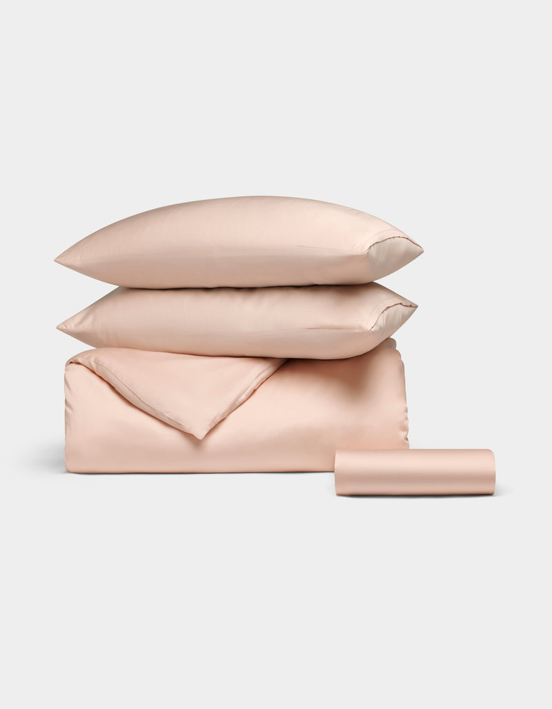 The Bamboo Bedding Essential Bundle by Cozy Earth, featuring light pink satin pillows, a folded duvet, and a rolled sheet, is neatly arranged against a light grey background.
