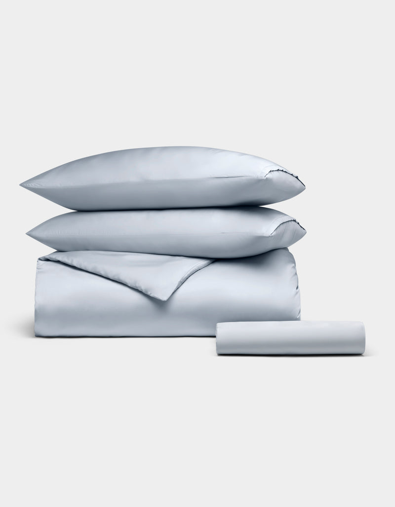 The Cozy Earth Bamboo Bedding Essential Bundle, featuring two pillows, a folded duvet, and a rolled sheet in light gray, is neatly stacked and displayed against a plain white background.