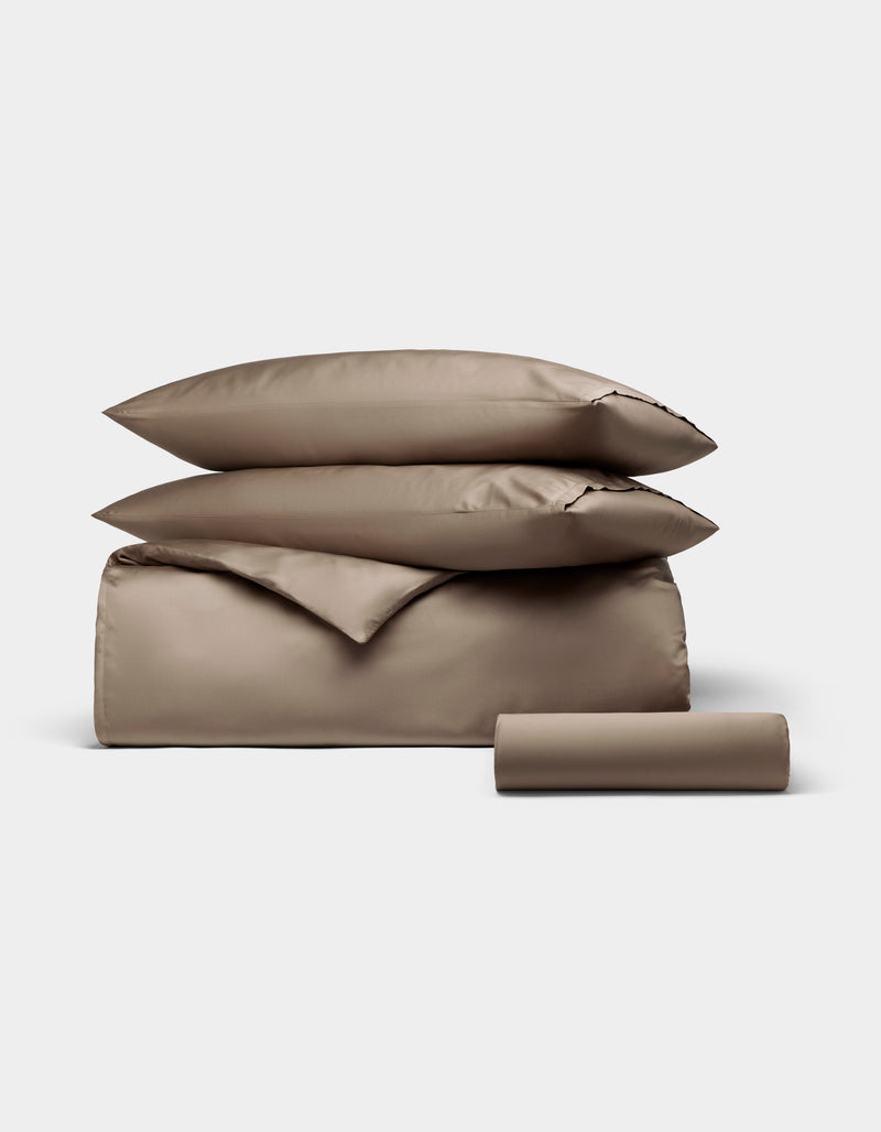 The Cozy Earth Bamboo Bedding Essential Bundle, featuring two pillows, a duvet cover, and a cylindrical bolster in taupe, is neatly folded and displayed against a plain light gray background.