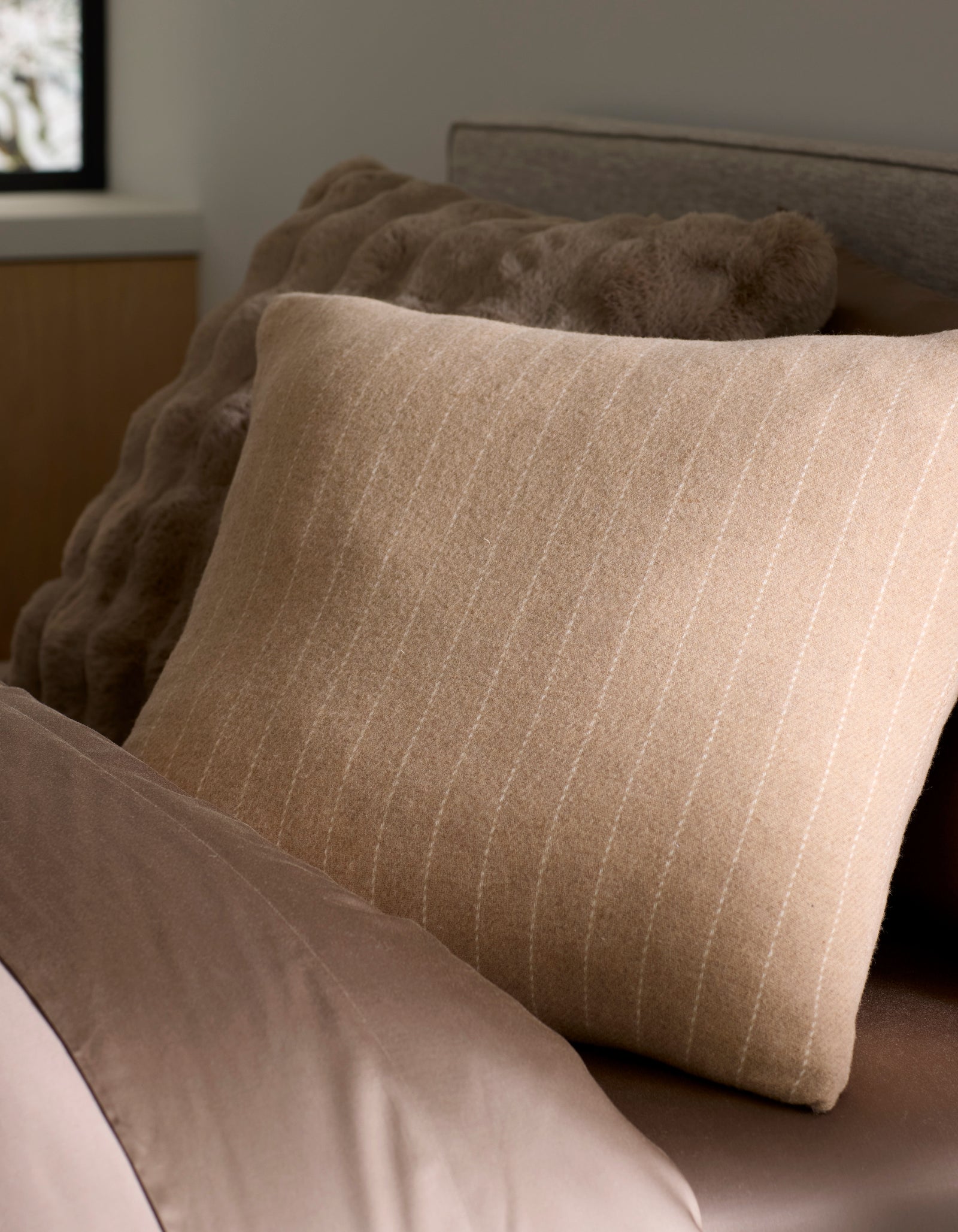 A close-up of a neatly made bed features Cozy Earth's Florence Pillow, a beige striped accent placed in front of a textured brown cushion. Soft brown bedding and a window with blurred outdoor light complete the cozy scene. 