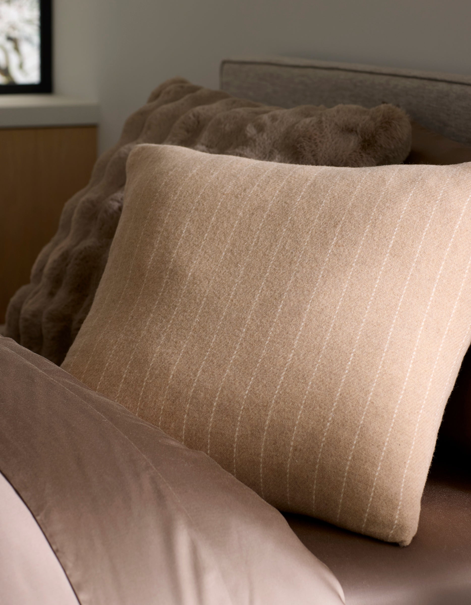 A close-up of a neatly made bed features Cozy Earth's Florence Pillow, a beige striped accent placed in front of a textured brown cushion. Soft brown bedding and a window with blurred outdoor light complete the cozy scene. 