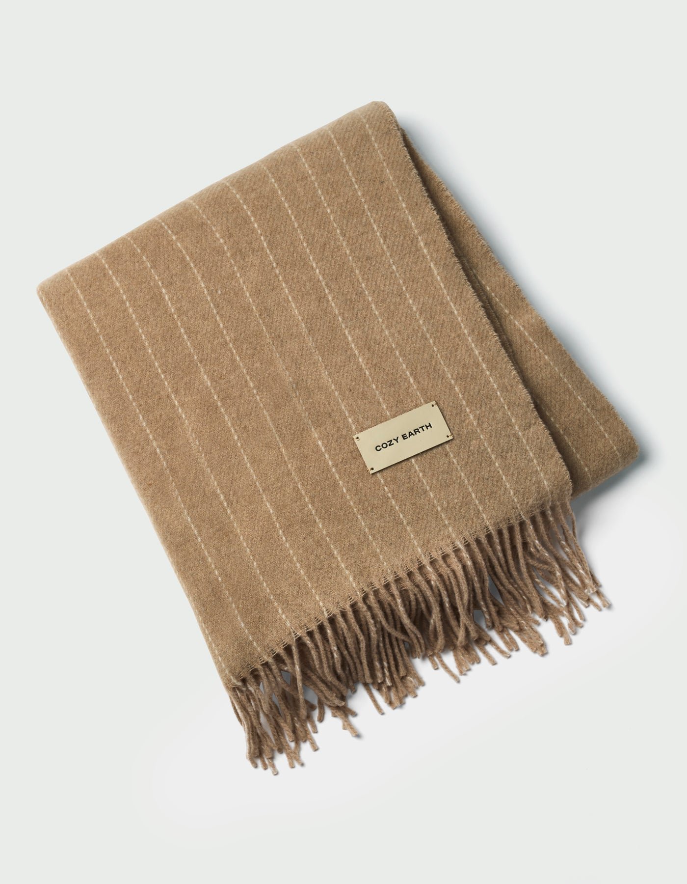 The Florence Throw by Cozy Earth is a tan, folded blanket with white pinstripes, fringed edges, and a rectangular "COZY EARTH" label on top, shown against a white background.|Color:Brandy