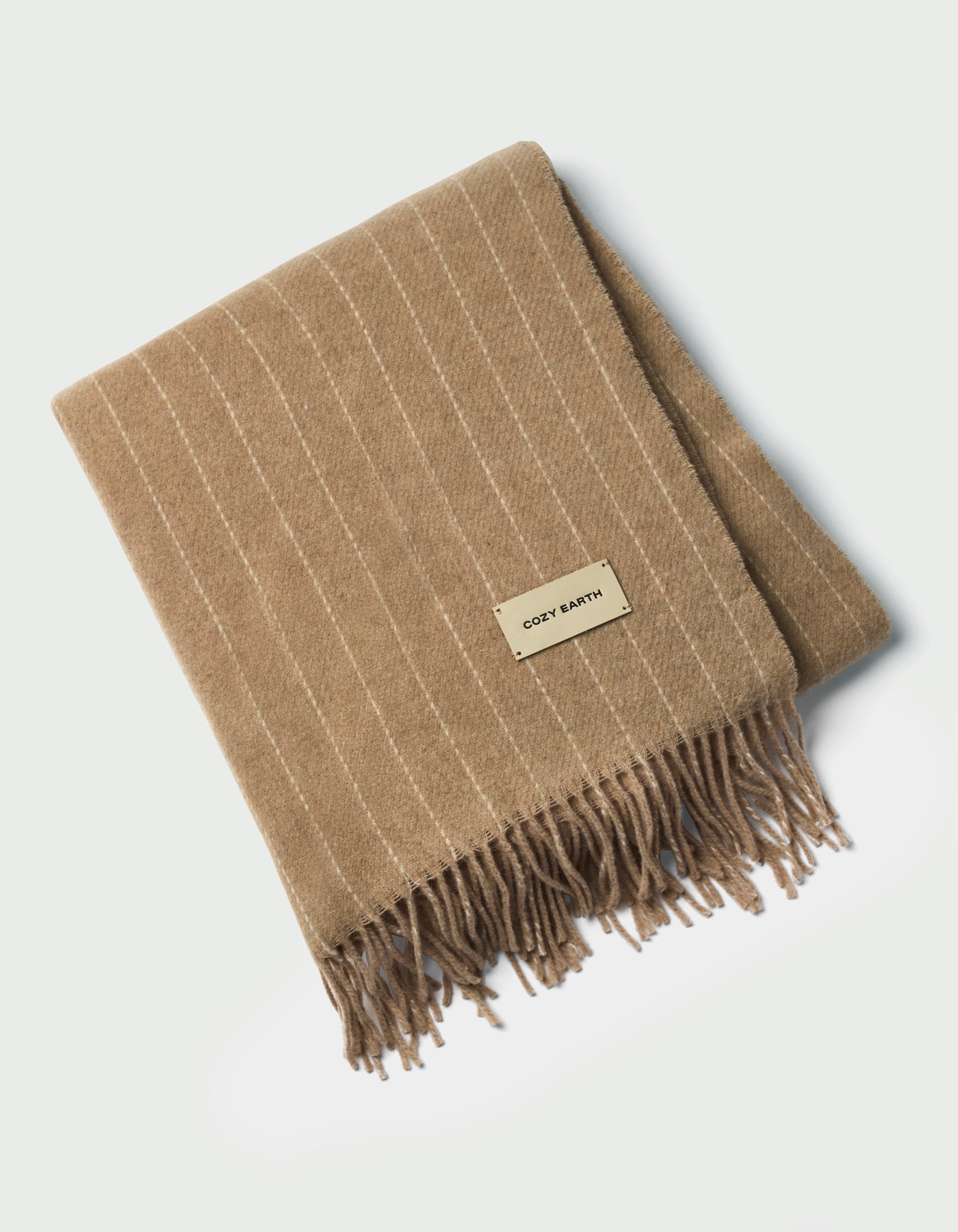 The Florence Throw by Cozy Earth is a tan, folded blanket with white pinstripes, fringed edges, and a rectangular "COZY EARTH" label on top, shown against a white background.