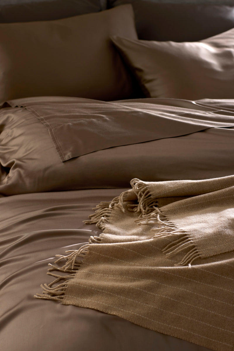 A neatly made bed with brown sheets, matching pillowcases, and the Cozy Earth Florence Throw in beige, featuring a fringed edge and folded at the corner. The bedding creates a smooth, inviting, and warm atmosphere.