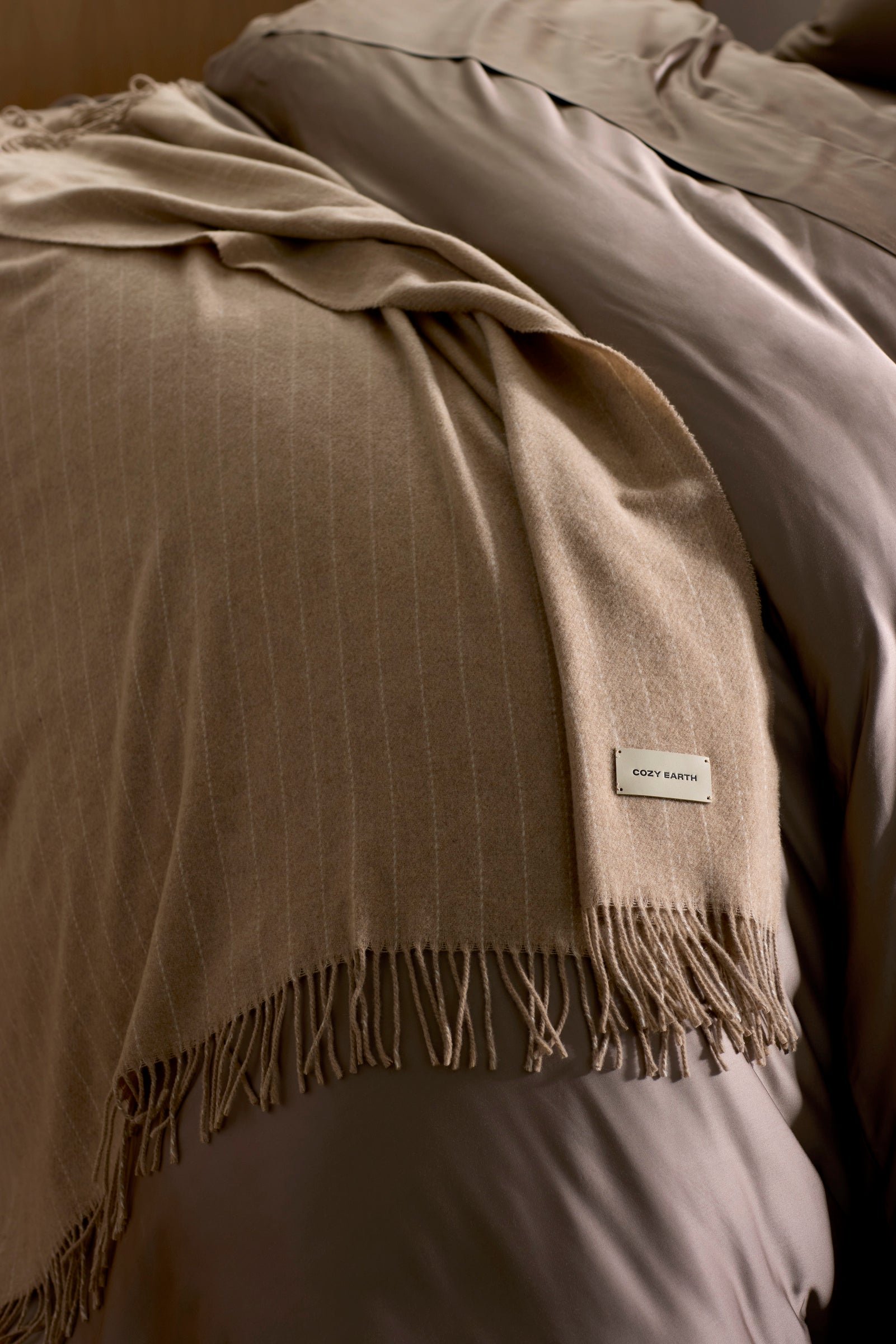 The Florence Throw by Cozy Earth, a soft beige blanket with fringed edges and a "COZY EARTH" label, is draped over taupe bedding on a neatly made bed.