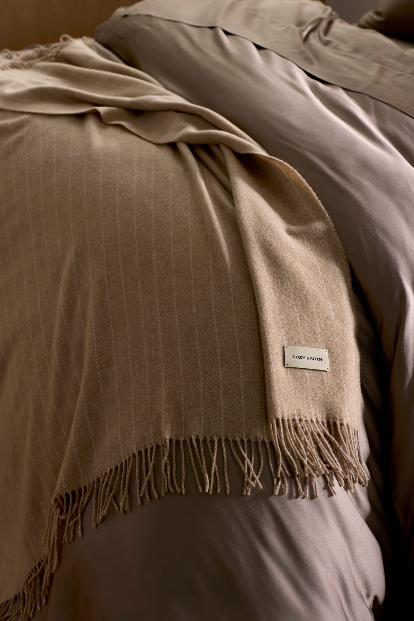 The Florence Throw by Cozy Earth, a soft beige blanket with fringed edges and a "COZY EARTH" label, is draped over taupe bedding on a neatly made bed.