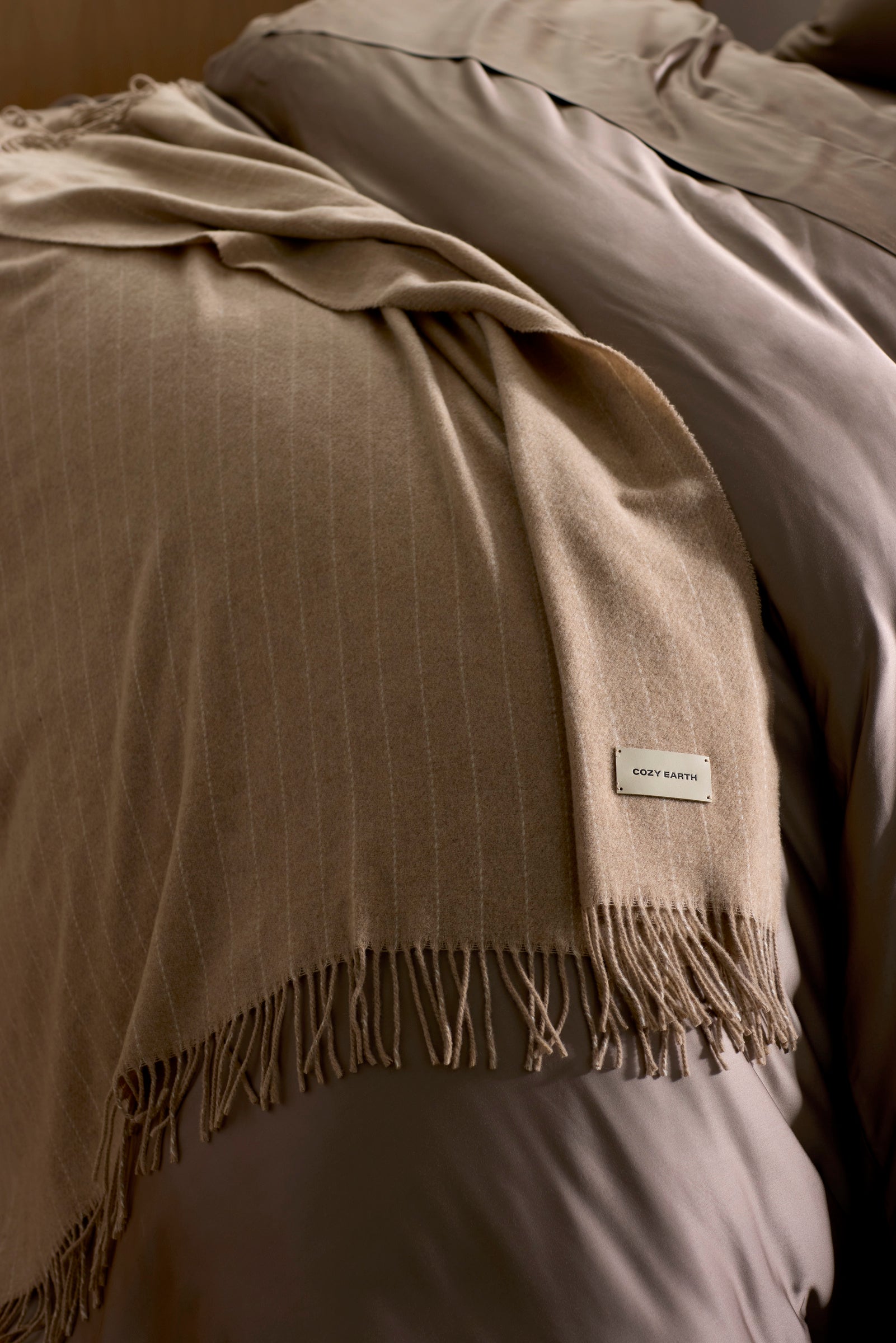 The Florence Throw by Cozy Earth, a soft beige blanket with fringed edges and a "COZY EARTH" label, is draped over taupe bedding on a neatly made bed.|Color:Brandy