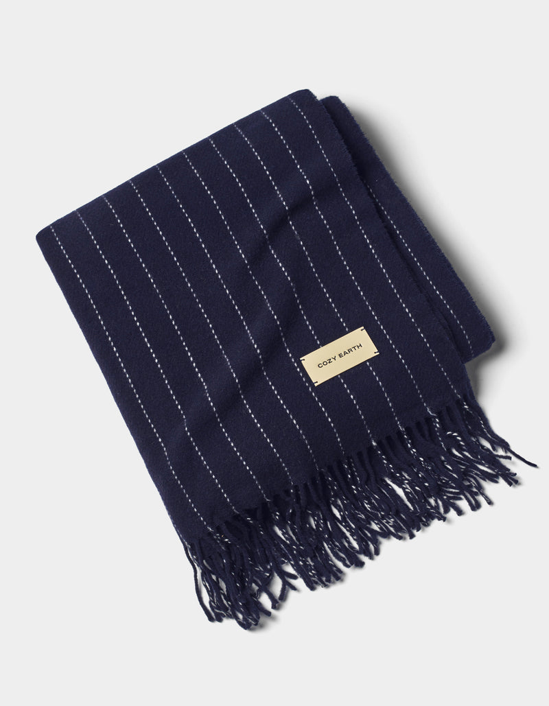 A neatly folded Florence Throw by Cozy Earth, featuring navy blue with thin white vertical stripes, fringed edges, and a beige fabric label on one end, displayed against a light background.