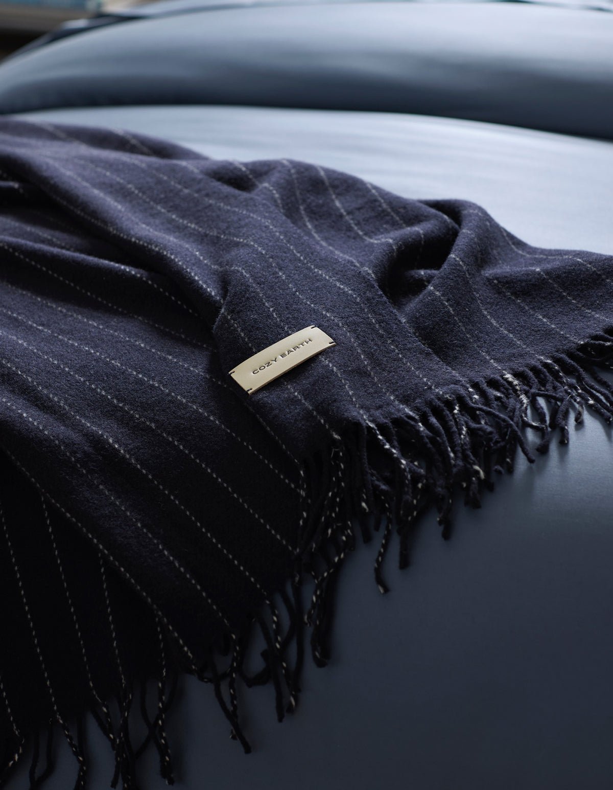 A dark blue, pinstriped Florence Throw by Cozy Earth with fringe is draped over a smooth, light blue surface. A rectangular label showing "COZY EARTH" is attached to the blanket. 