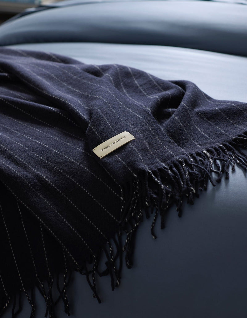 A dark blue, pinstriped Florence Throw by Cozy Earth with fringe is draped over a smooth, light blue surface. A rectangular label showing "COZY EARTH" is attached to the blanket.