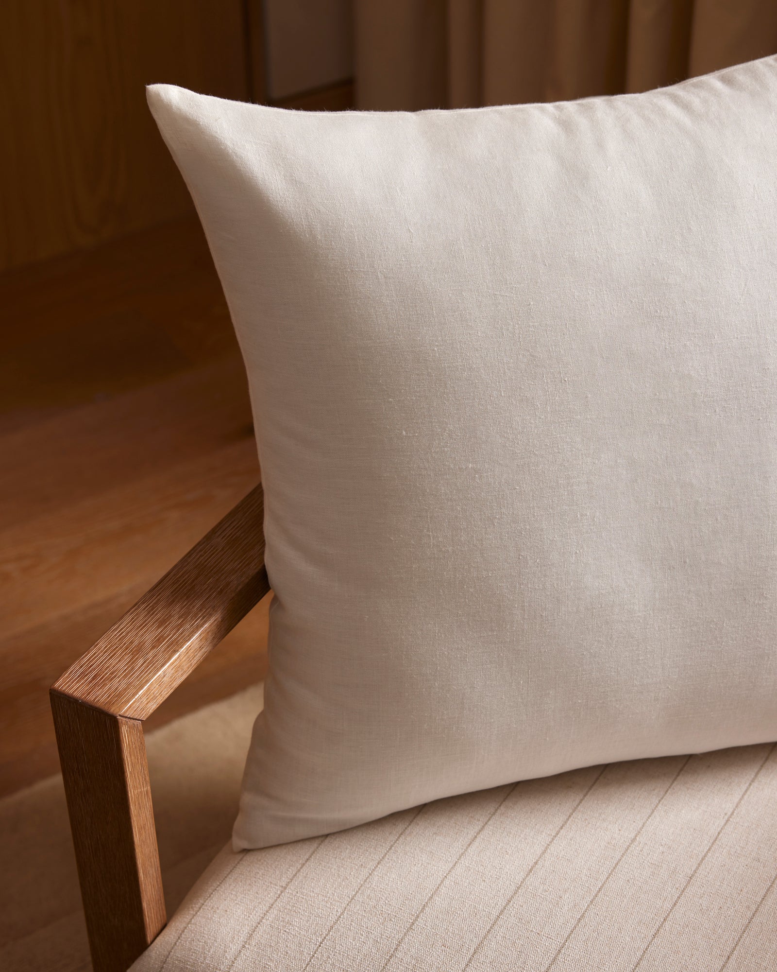 A close-up of Cozy Earth’s Linen Throw Pillow in light beige, placed on a wooden chair with a textured cushion. The scene is warm and minimalistic, featuring soft lighting and wooden flooring in the background.
