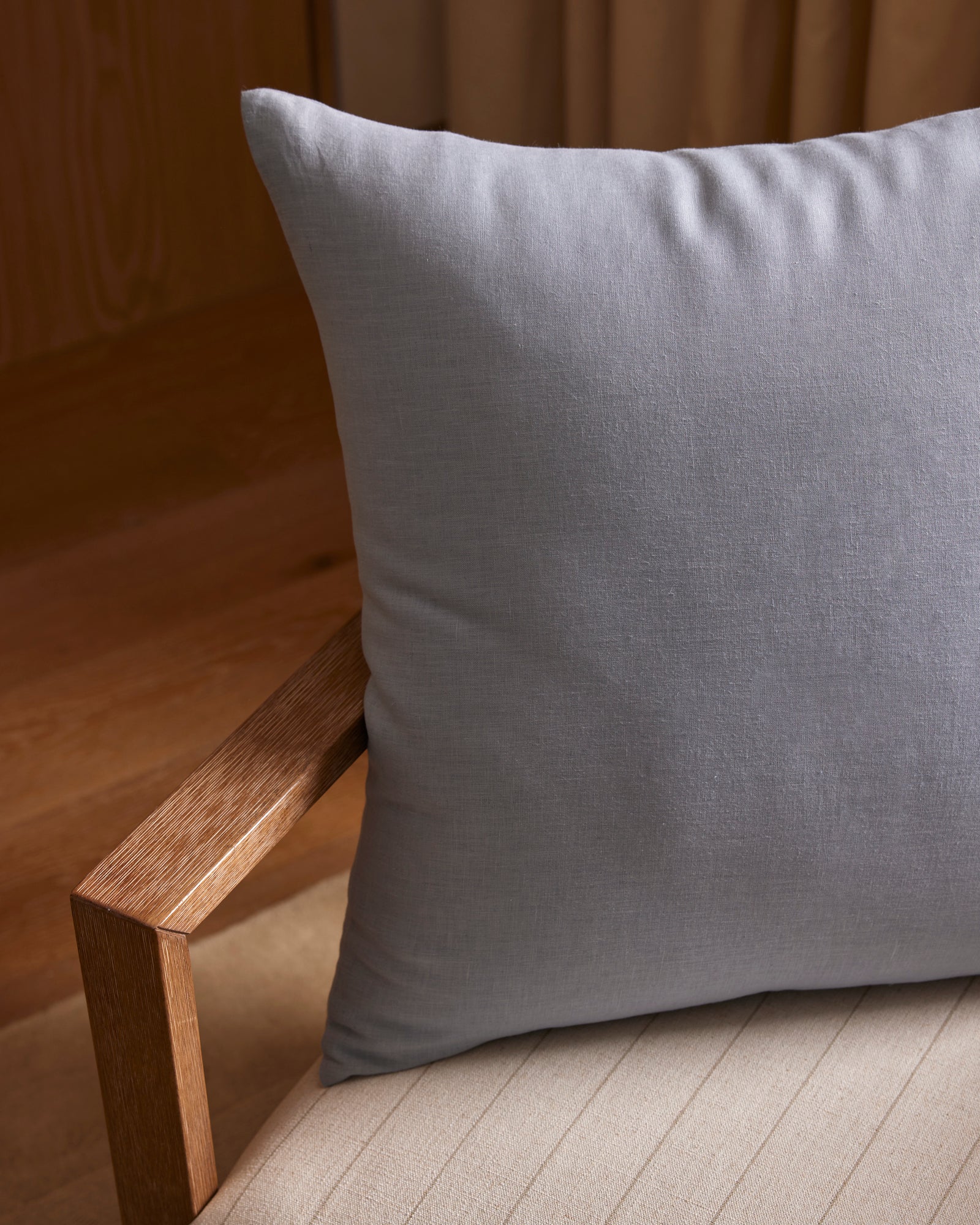 The Cozy Earth Linen Throw Pillow, in light gray, sits on a beige upholstered chair with a wooden armrest, set against a backdrop featuring wood accents and curtains.