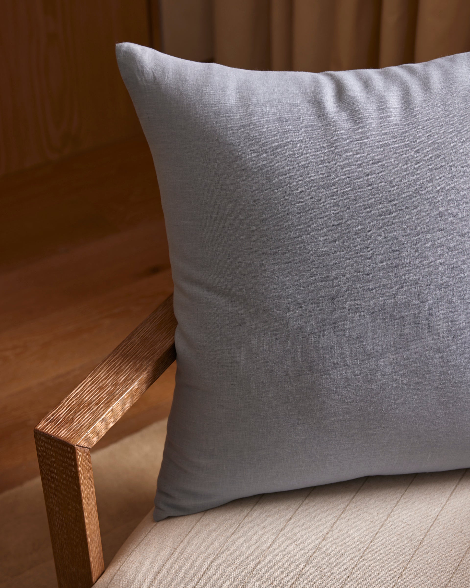 A Cozy Earth Linen Throw Pillow in light gray sits on a beige cushioned chair with wooden arms, placed in a warmly lit room featuring wooden flooring and tan curtains.