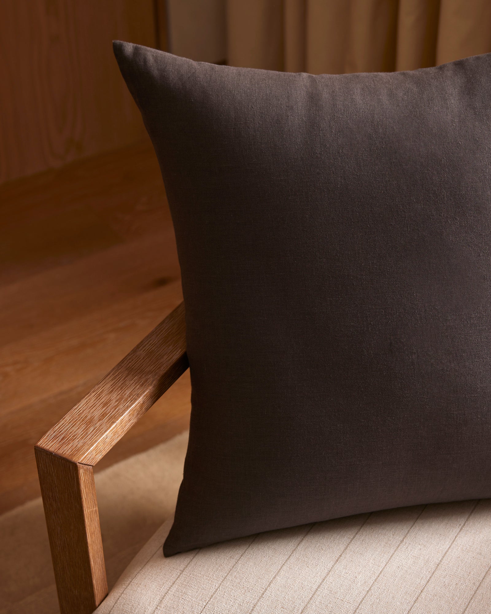 A close-up of the Cozy Earth Linen Throw Pillow in dark brown, placed on a light cushioned chair with a wooden armrest, set in a softly lit room featuring wooden flooring and beige curtains.