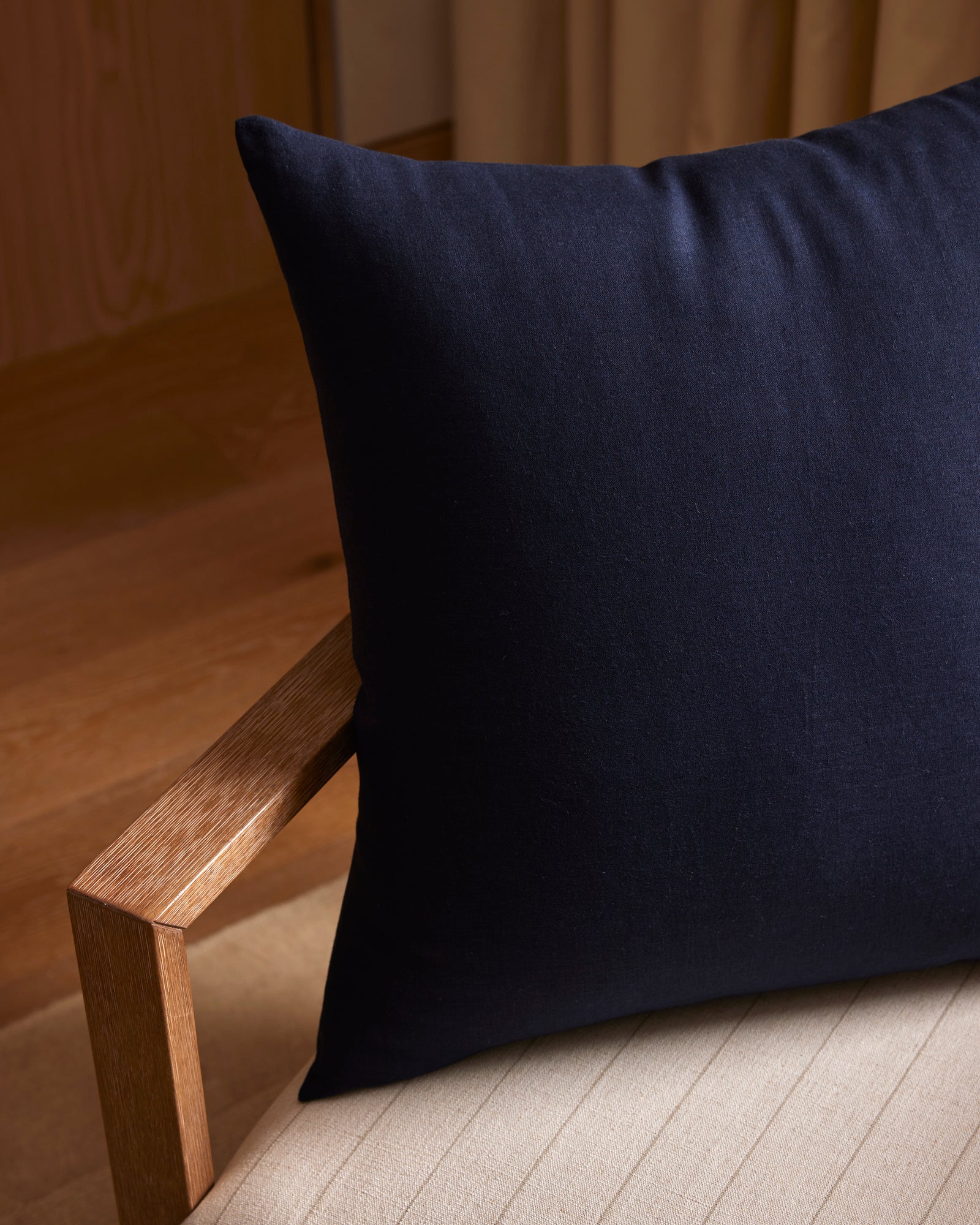A close-up of the Cozy Earth Linen Throw Pillow in dark navy blue rests on a light upholstered chair with wooden armrests, set against wooden flooring and beige curtains.