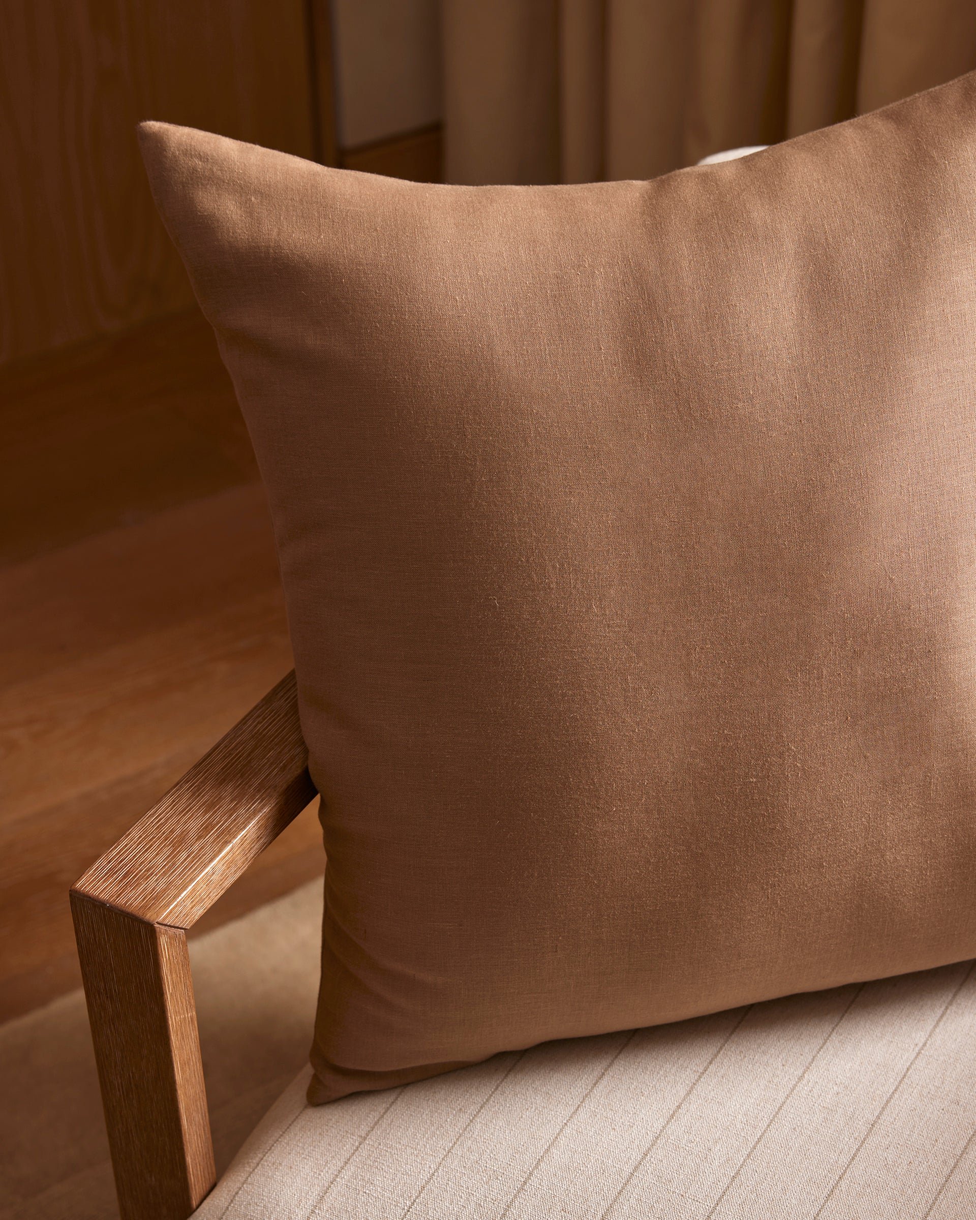 A Cozy Earth Linen Throw Pillow sits on a light beige upholstered chair with a wooden armrest, in a softly lit room featuring wooden flooring and neutral curtains.|Color:Maple