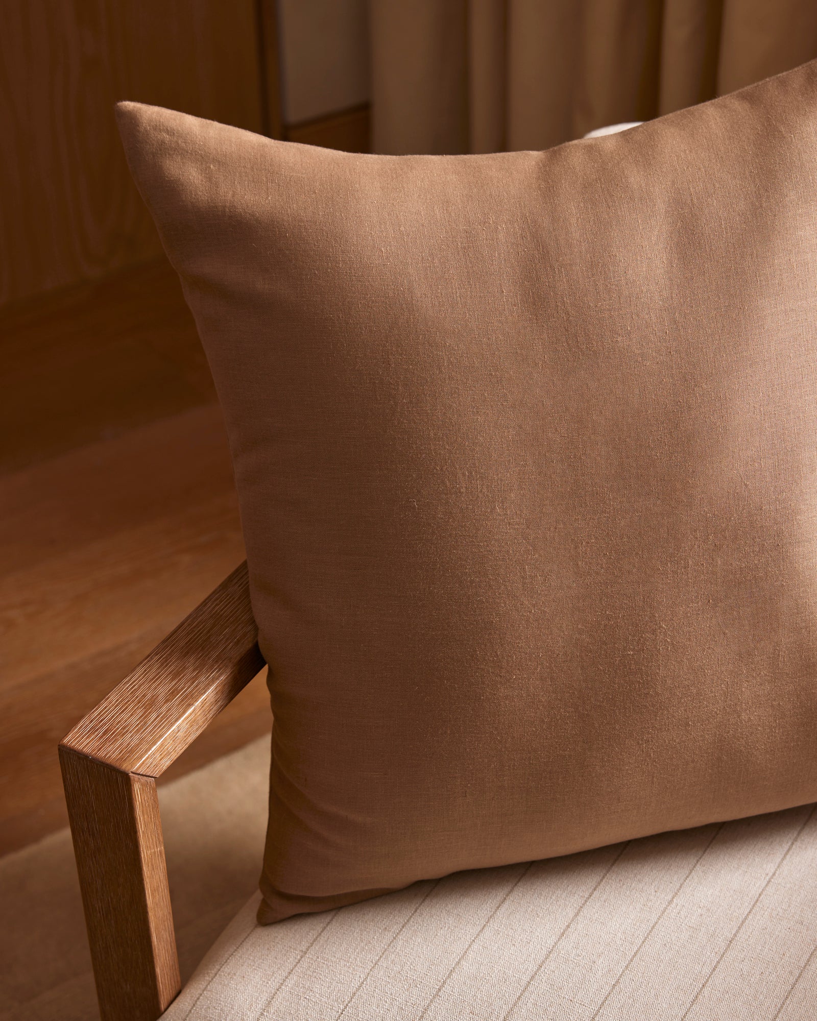 A Cozy Earth Linen Throw Pillow sits on a light beige upholstered chair with a wooden armrest, in a softly lit room featuring wooden flooring and neutral curtains.