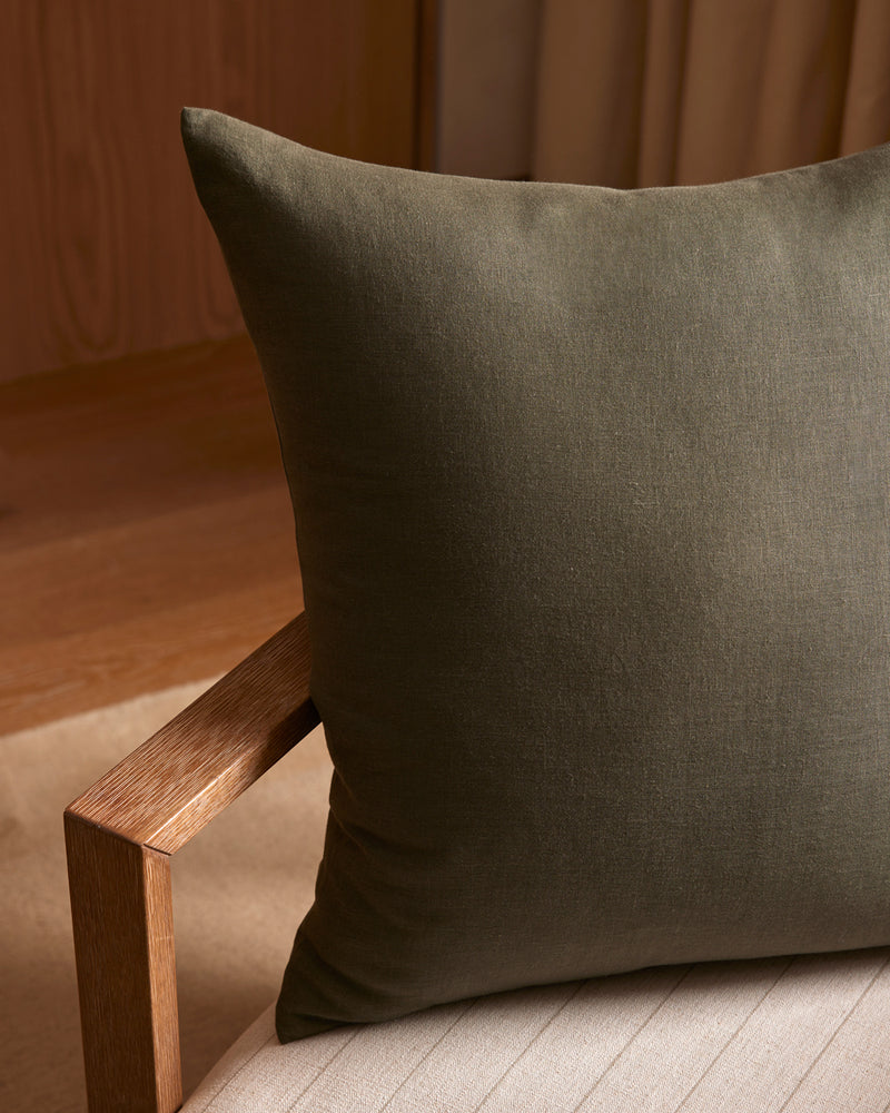 A close-up of the Cozy Earth Linen Throw Pillow in dark olive green rests on a wooden chair with a beige cushion, set against wood paneling and beige curtains.