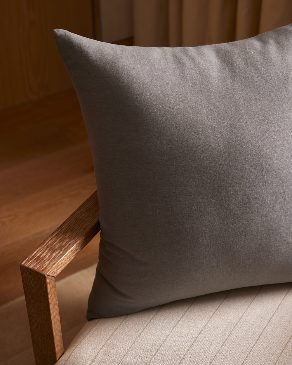 A Cozy Earth Linen Throw Pillow in gray sits on a light beige cushioned chair with a wooden armrest, set in a softly lit room featuring wooden floors and curtains in the background.