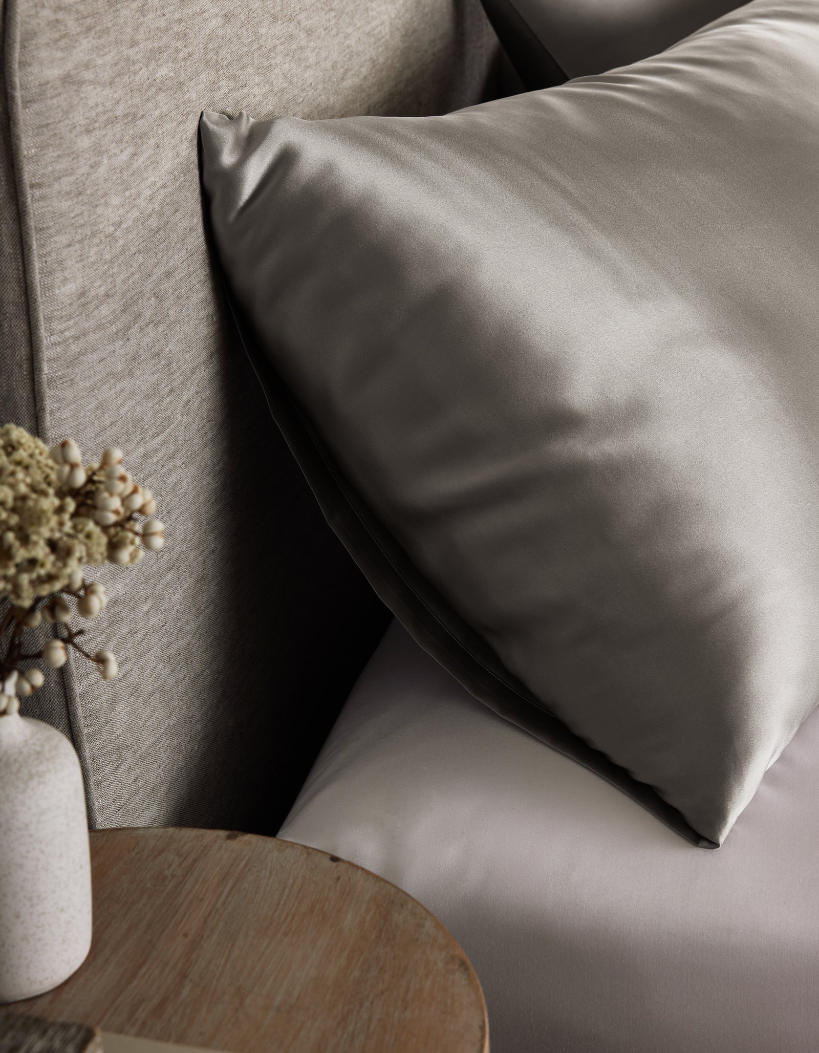 A close-up of a neatly made bed features the Cozy Earth Silk Pillowcase in smooth, shiny gray beside a small wooden table topped with a vase of dried flowers.