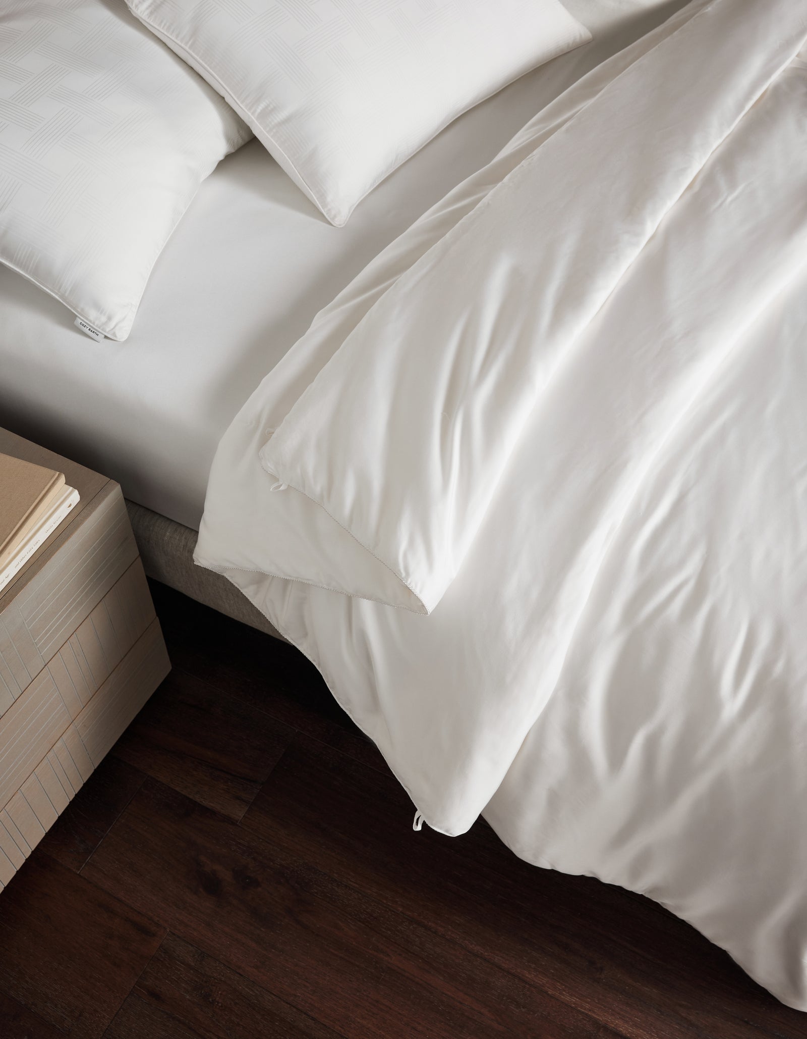 A bed with crisp white sheets, pillows, and a Cozy Earth Silk Comforter sits beside a wooden nightstand stacked with books on a dark wooden floor.