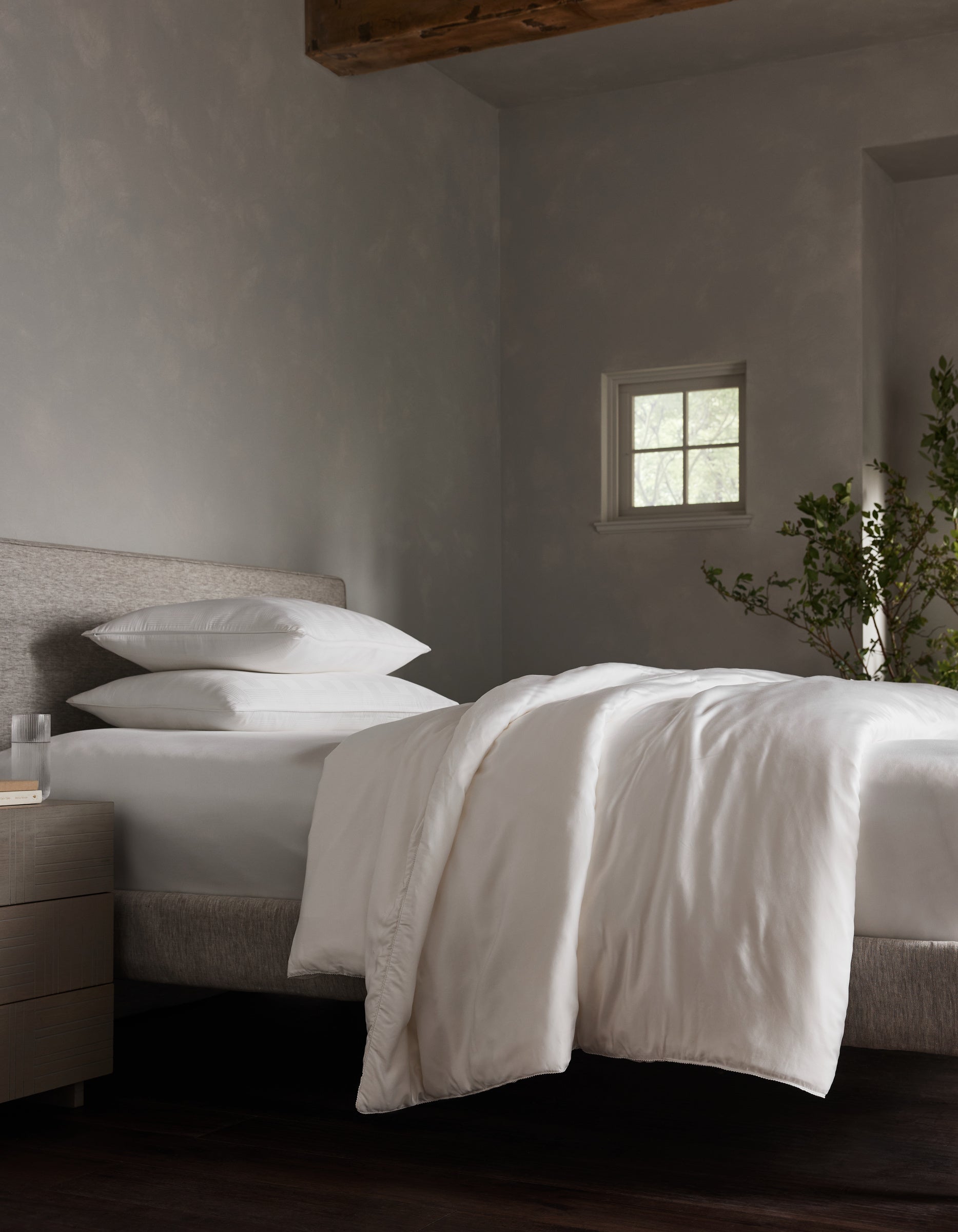 A neatly made bed features Cozy Earth's Silk Comforter with crisp white bedding and pillows, beside a small nightstand holding a glass of water. A small window and green plant add to the softly lit, minimalist bedroom atmosphere.|Filling:Standard