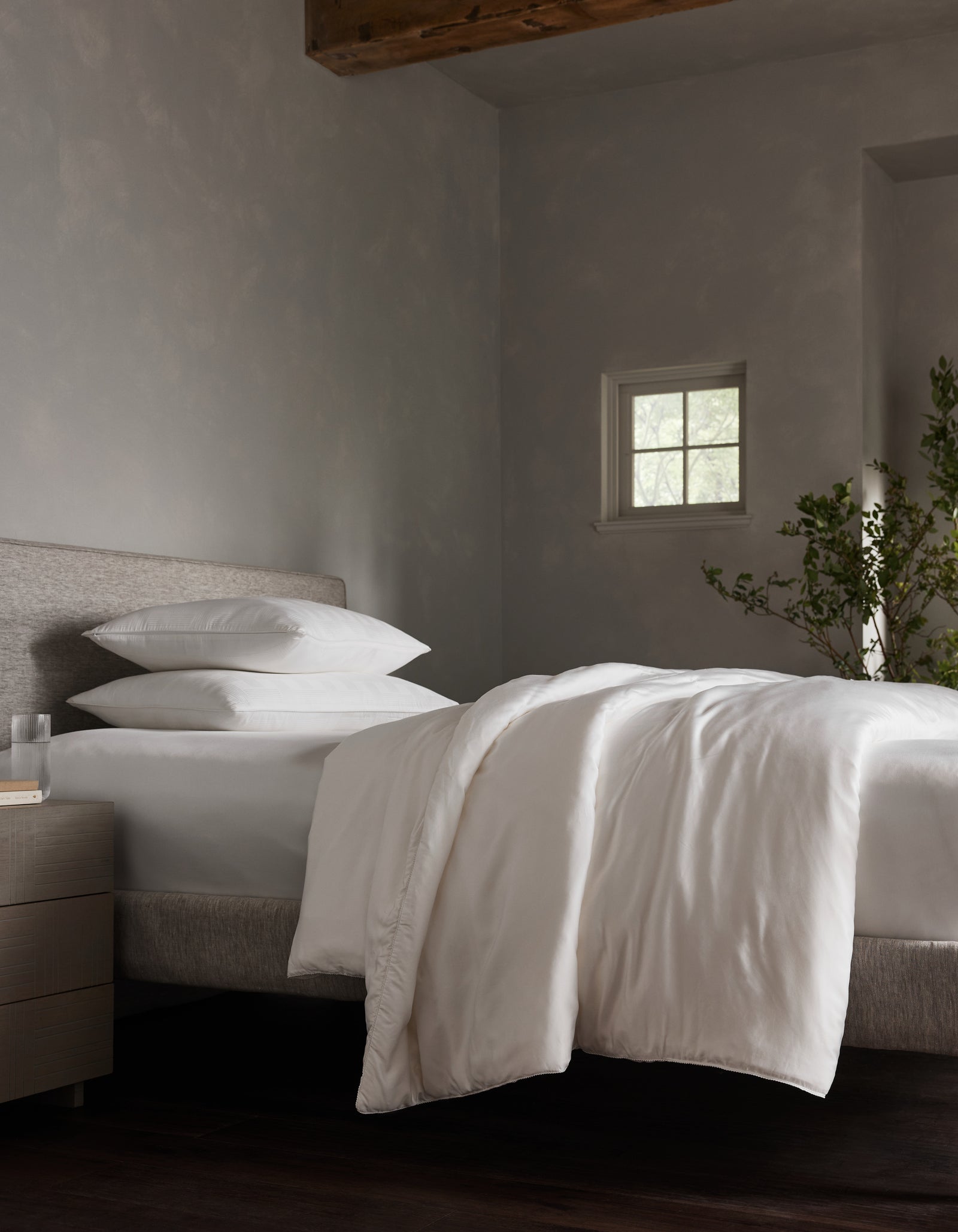 A minimalist bedroom with a neatly made bed features Cozy Earth’s Silk Comforter in white. Sunlight filters through a small window, illuminating the space with neutral tones, a bedside plant, and a nightstand holding water and a glass.