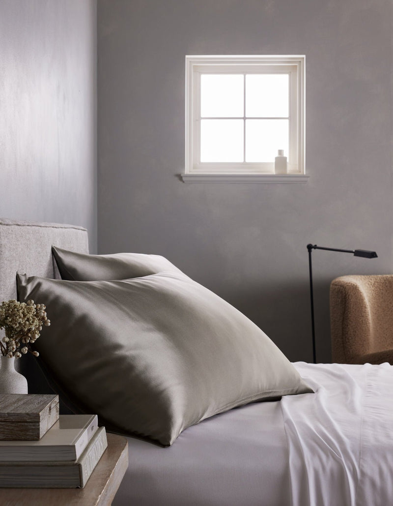 A minimalist gray bedroom features a neatly made bed with large Cozy Earth Silk Pillowcases in gray and white sheets, set beside a small stack of books, dried flowers, a beige chair, and a window that lets in natural light.